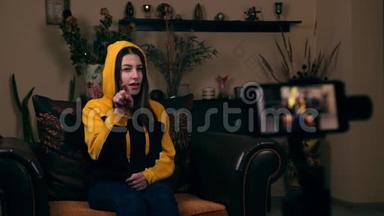 一位欧洲貌美的年轻女孩坐在家里的扶手椅上，在手机上录制视频视频，并大拇指
